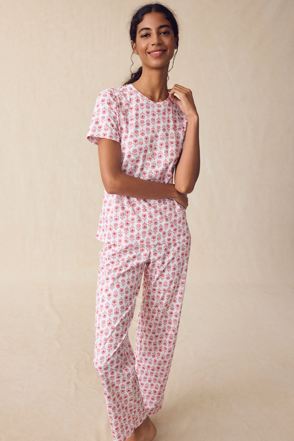 A person smiles, wearing the Pima Crew Short-Long Set in Rosewater Sprig—white pajamas with a pink floral block print, featuring a short-sleeve top and high-waisted pants—set against a beige background.