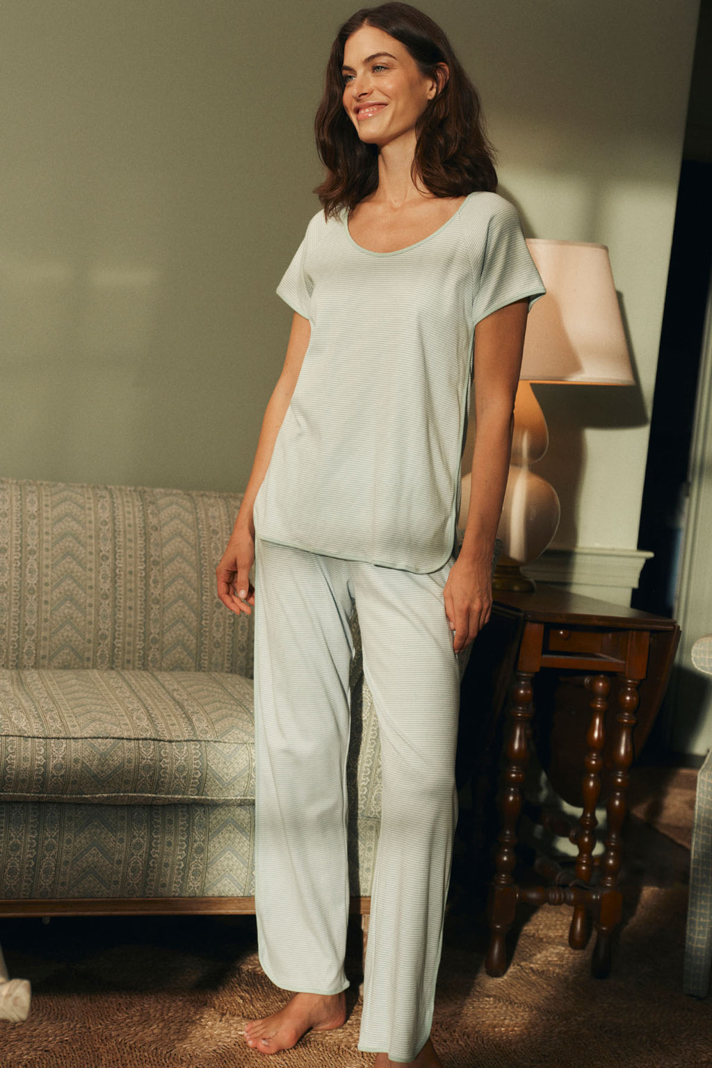 A woman smiles in a softly lit room, wearing the Pima Bundle in Parisian Green—light-colored, short-sleeved Pima cotton pajamas with an elastic waistband. She stands beside a striped sofa, with a lamp and wooden side table behind her.