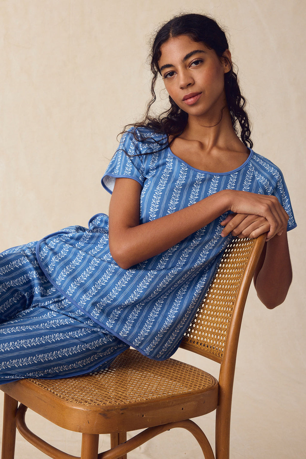 A woman sits sideways on a wooden chair, confidently facing the camera in the Pima Bundle in Seaport Jasmine Vine—matching blue top and pants with white leaf patterns—set against a neutral background.