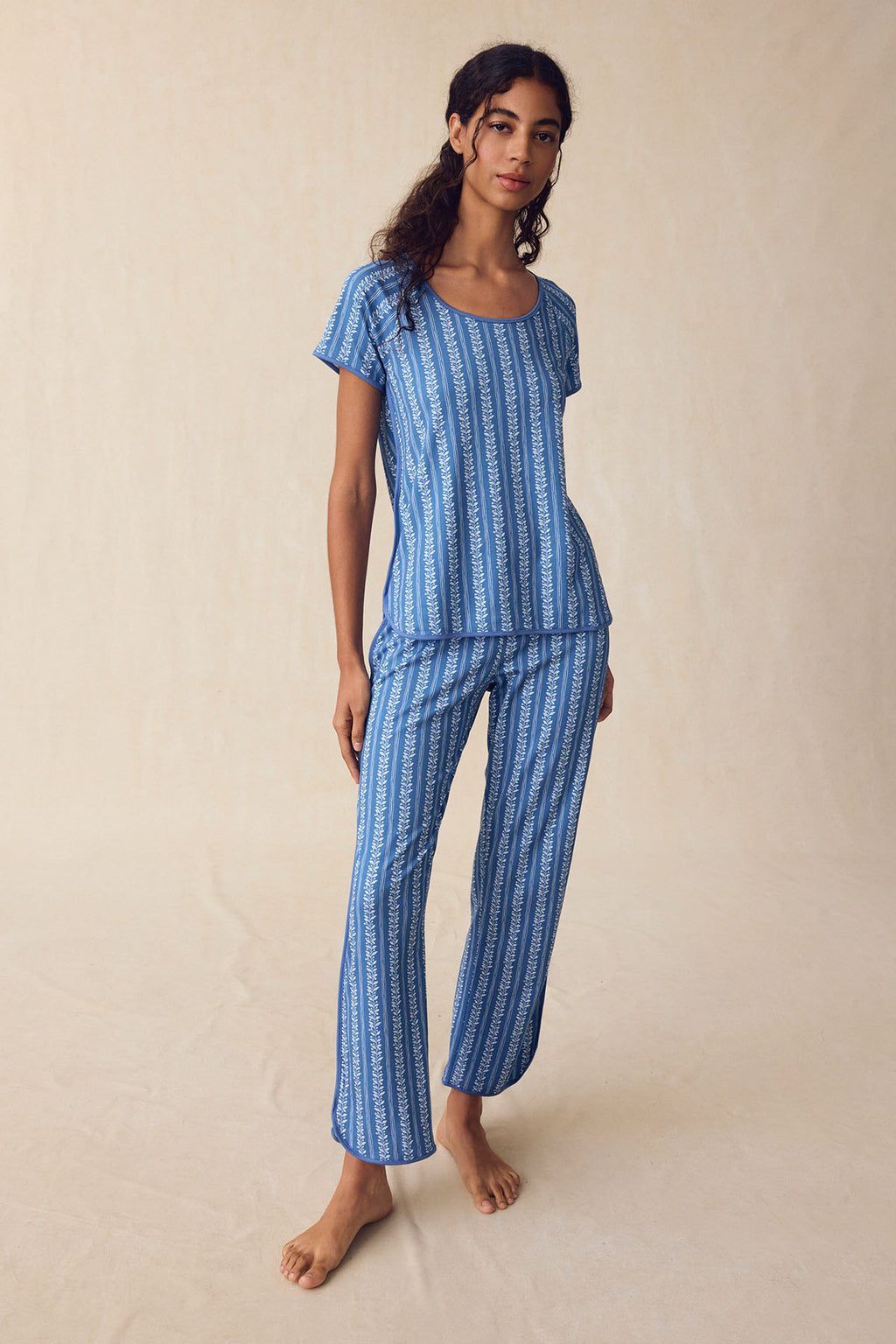 A woman stands against a plain background, wearing the Pima Bundle in Seaport Jasmine Vine—a matching blue short-sleeve pajama top and pants with a vertical jasmine vine print, crafted from soft Pima cotton.
