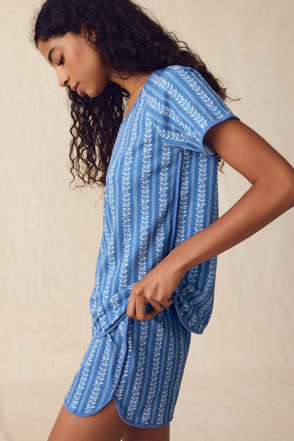 A woman in the Pima Bundle in Seaport Jasmine Vine—a blue, short-sleeved Pima cotton pajama set with a white floral pattern—stands in profile against a neutral background.