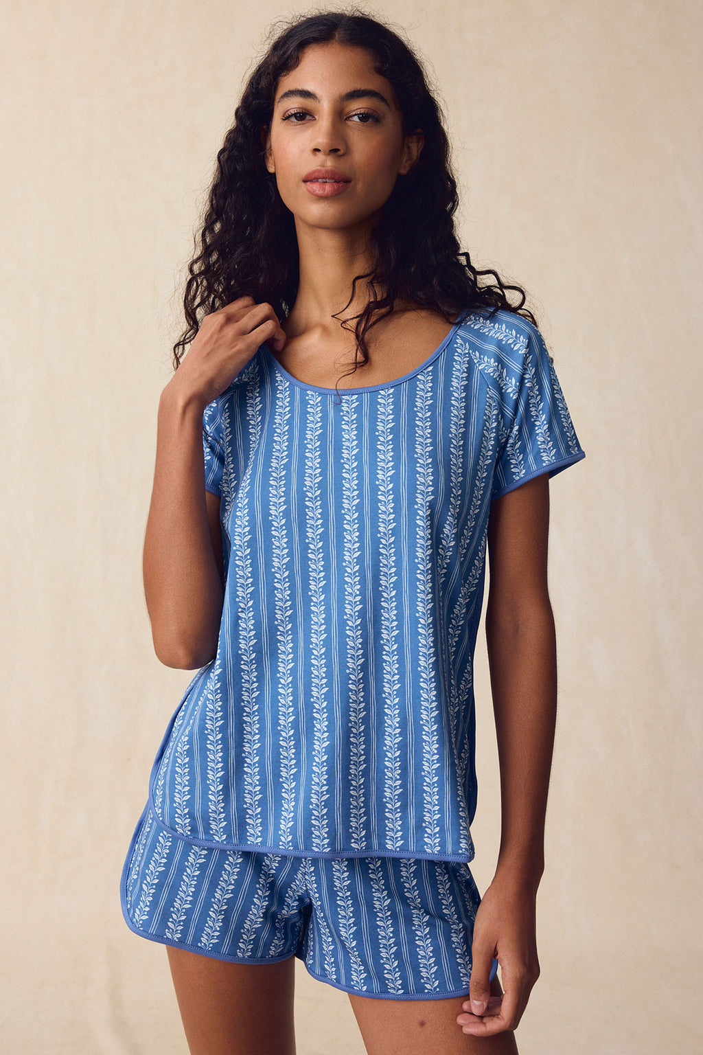 A woman wears the Pima Bundle in Seaport Jasmine Vine—a short-sleeved blue pajama set with vertical white vine stripes—standing before a neutral beige background.