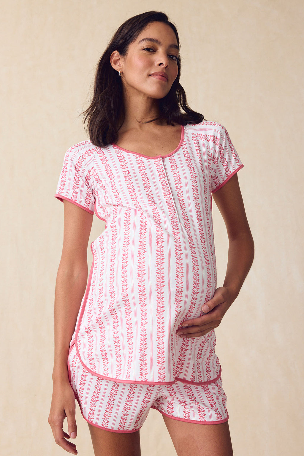 A pregnant woman wears the Pima Maternity Shorts Set in Winterberry Jasmine Vine, featuring pink and white floral stripes, as she softly smiles and holds her belly against a light beige background.