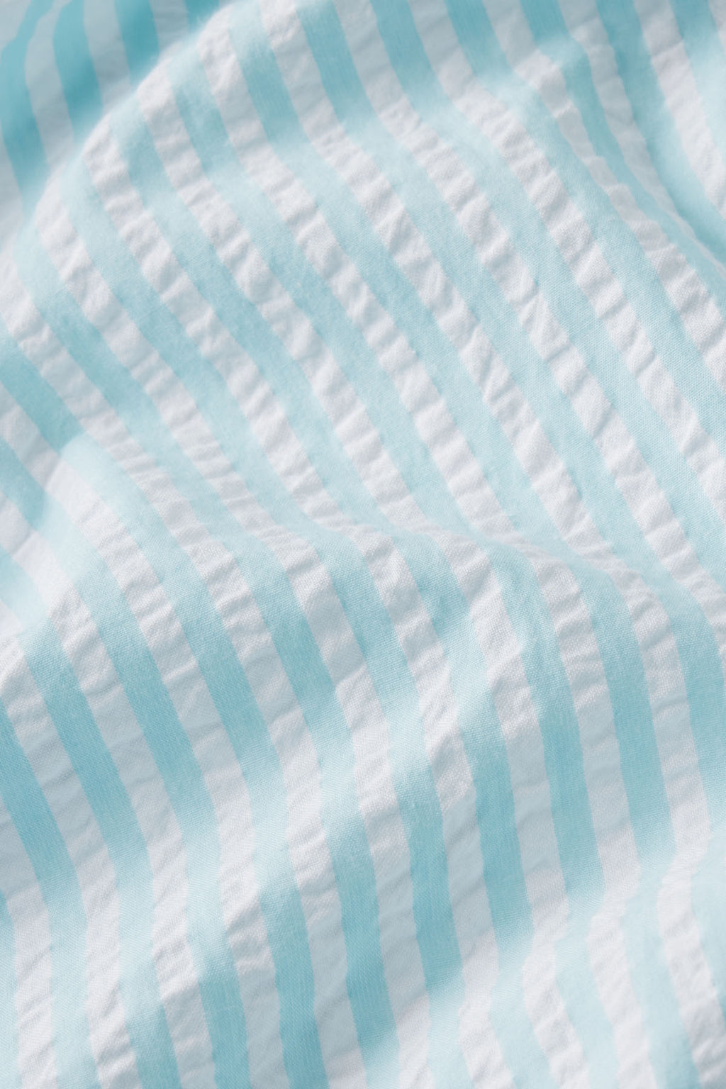 The Alice Nightgown in Salt Spray Seersucker features light blue and white waves, capturing a breezy feel with gentle folds and soft shadows, crafted from luxuriously soft Pima cotton.