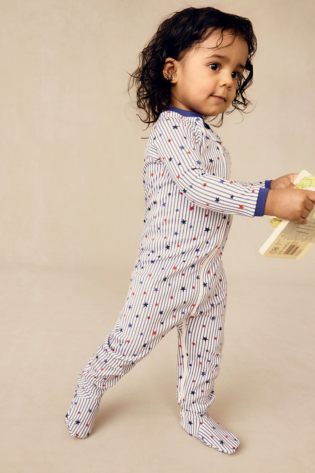 Toddler wearing white long-sleeved sleeper with red and blue stars and vertical stripes, blue cuffs at wrists and ankles.