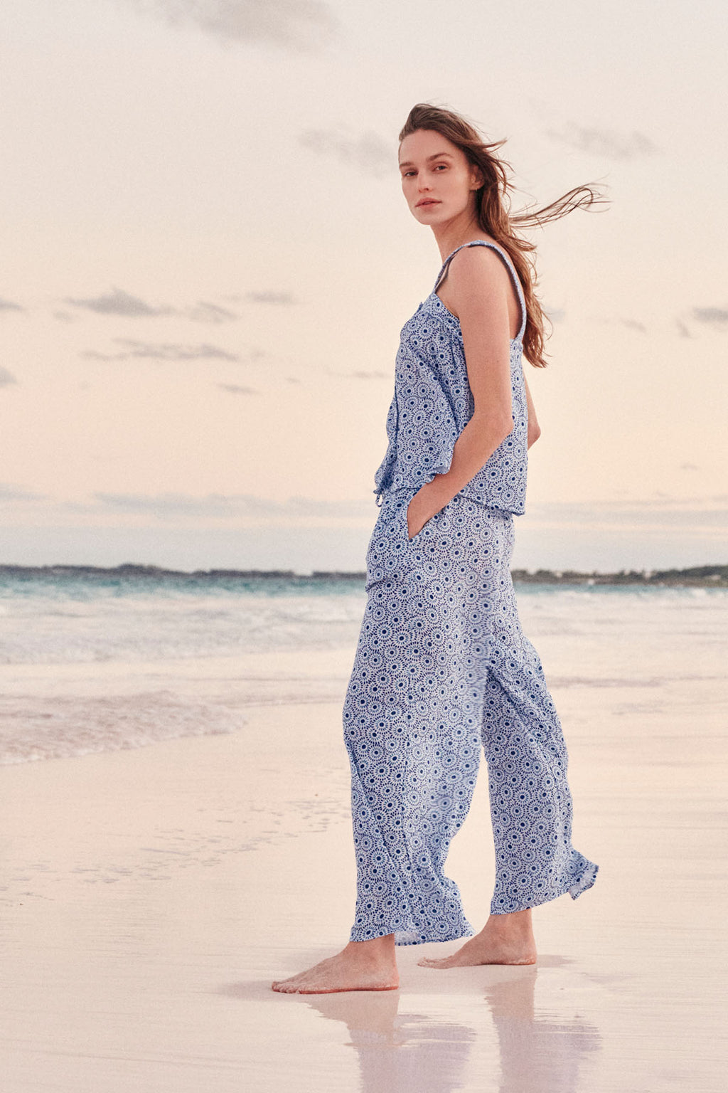A woman strolls along a sandy beach at sunset, wearing the Hamptons Pants Set in Lapis Dot Gauze, with gentle waves and a pastel sky in the background.