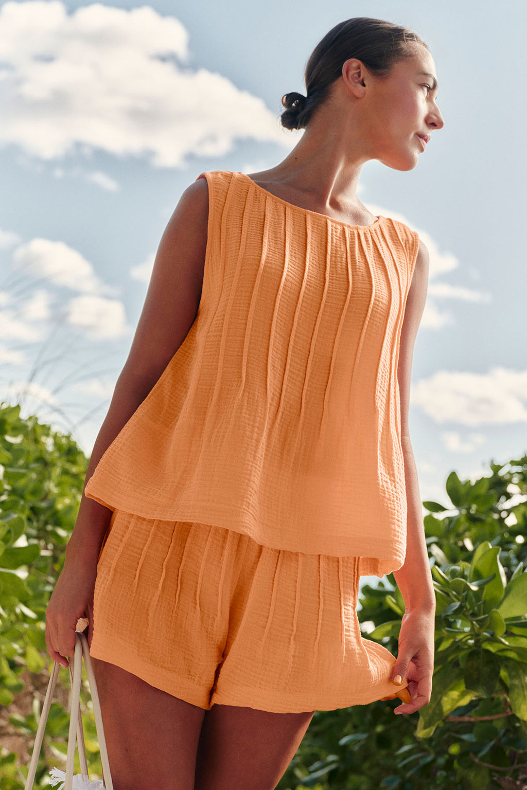 A woman stands outside beneath a partly cloudy sky, looking to the side, wearing the Monaco Shorts Set in Marigold Gauze—a loose-fitting, sleeveless top with matching cotton gauze shorts—surrounded by greenery.