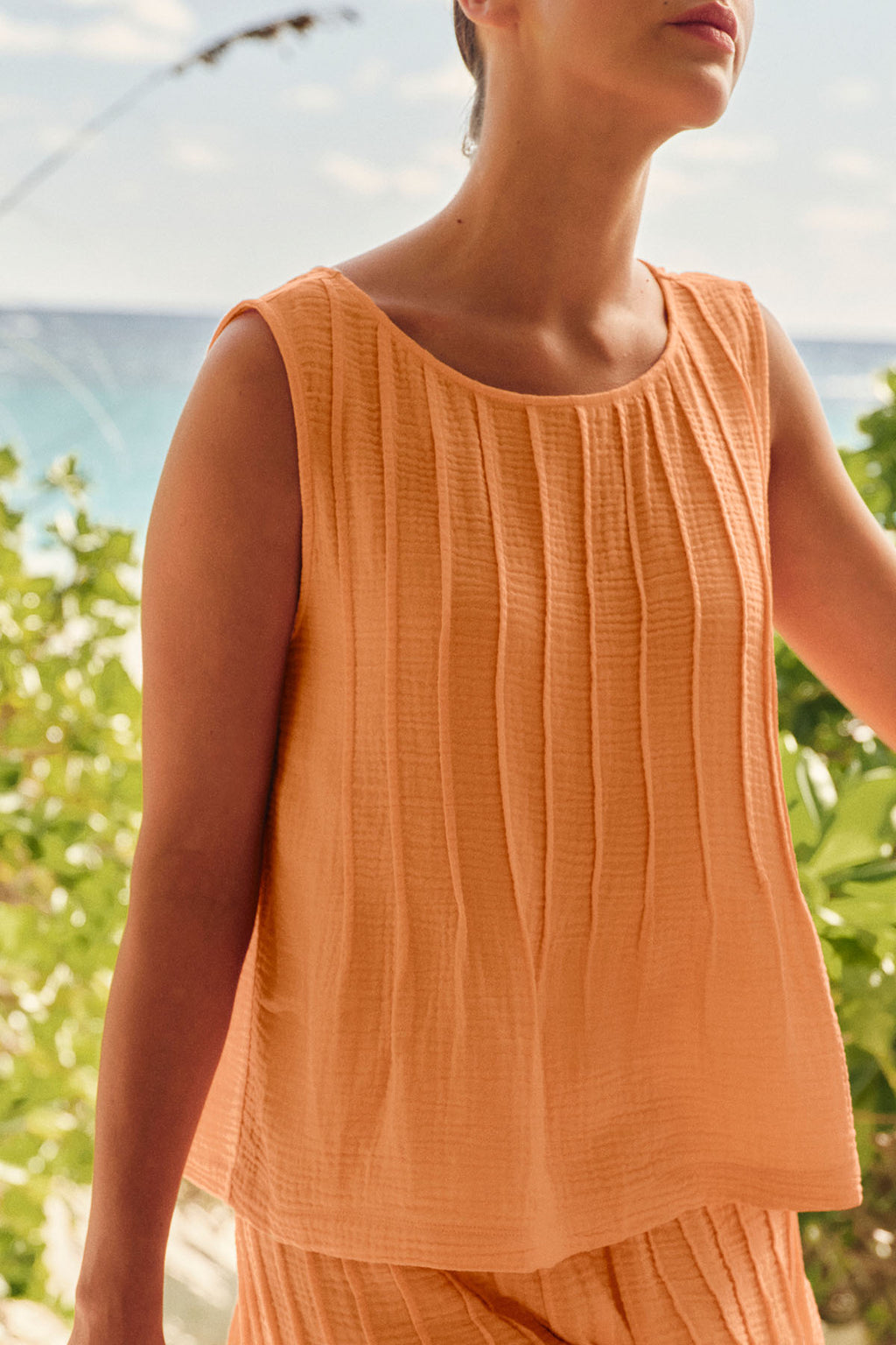 A person wears the Monaco Shorts Set in Marigold Gauze, standing outdoors with greenery and the ocean behind them. The scene is bright and summery.