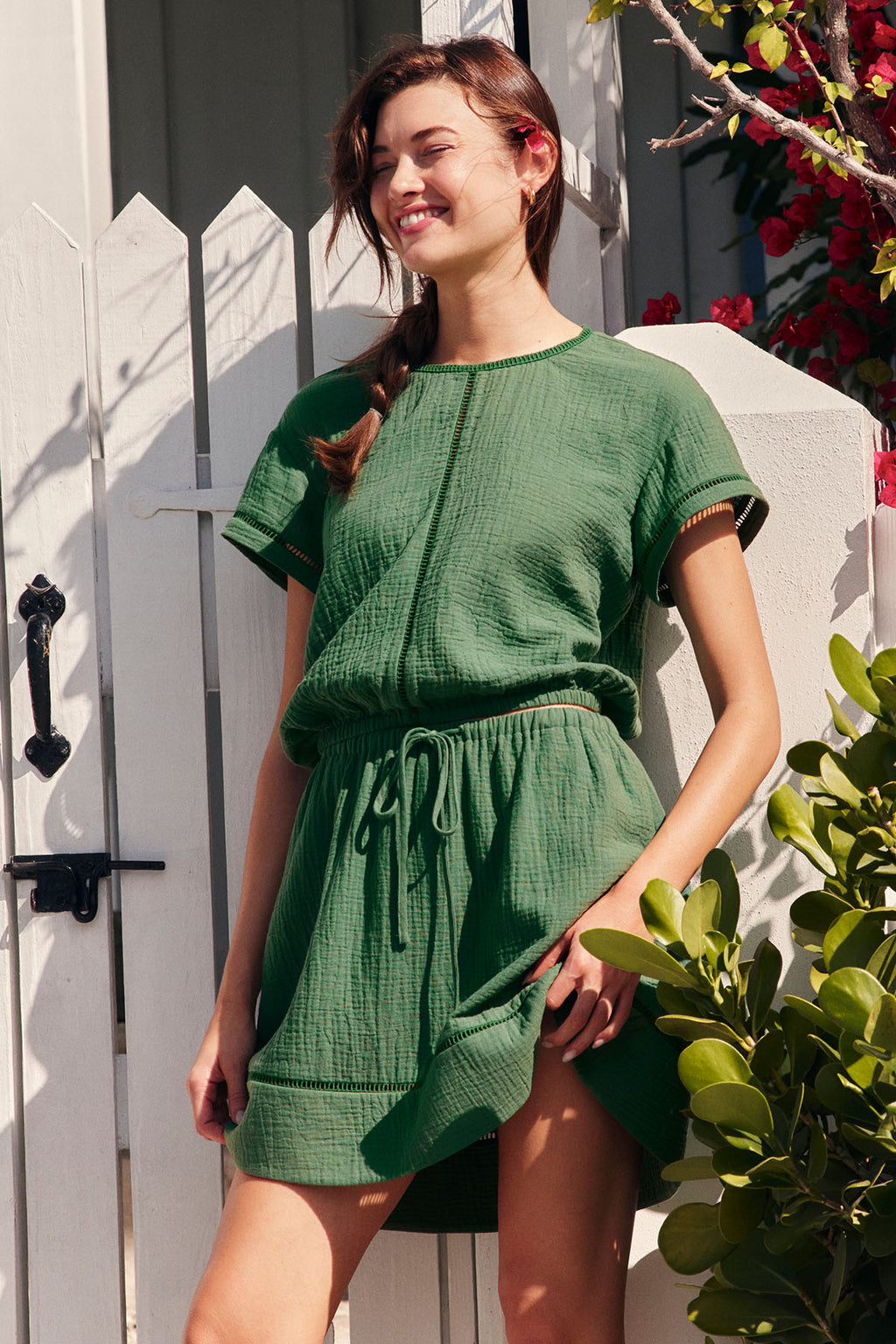 A smiling woman stands by a white gate, wearing the Cabana Skirt Set in Palm with a drawstring waist, surrounded by greenery and red flowers.
