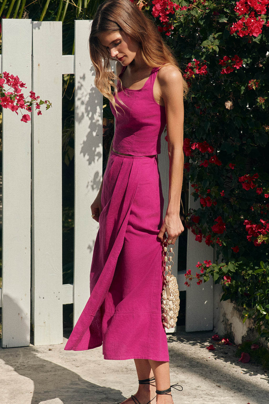 Wearing the Flute Skirt Set in Fuchsia, a woman stands outdoors by a white fence and vivid red flowers, holding a beaded handbag as sunlight creates soft shadows while she looks down.