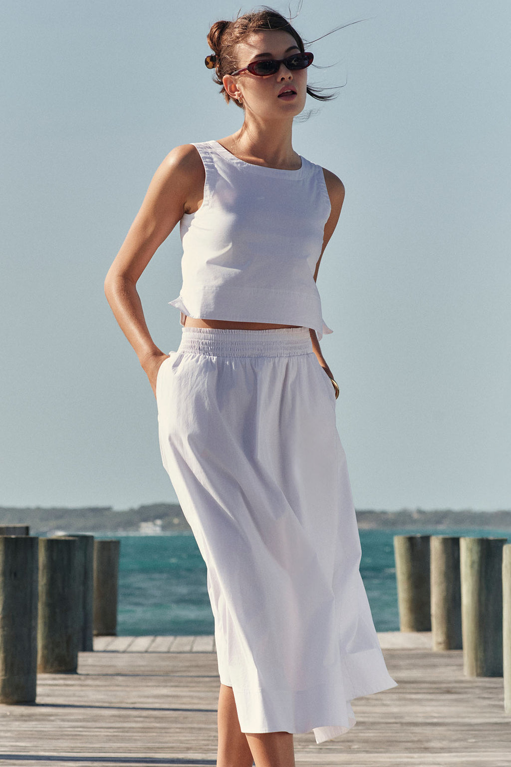 A woman in a sleeveless white top and the Terrace Skirt Set in White with a midi skirt stands on a wooden dock by the water, wearing sunglasses under a clear sky. The wrinkle-resistant cotton poplin adds style and comfort.