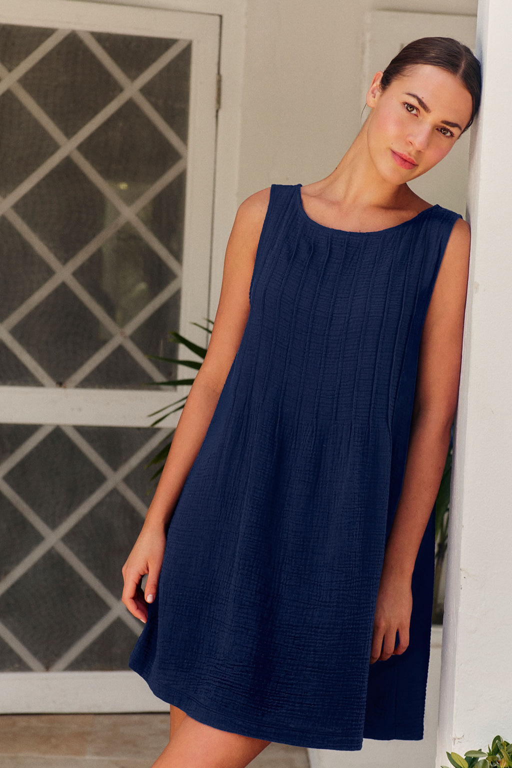A woman in a sleeveless, loose-fitting Monaco Nightgown in Blue Depths Gauze leans against a white wall near a crisscross-patterned screen door, gazing softly at the camera.