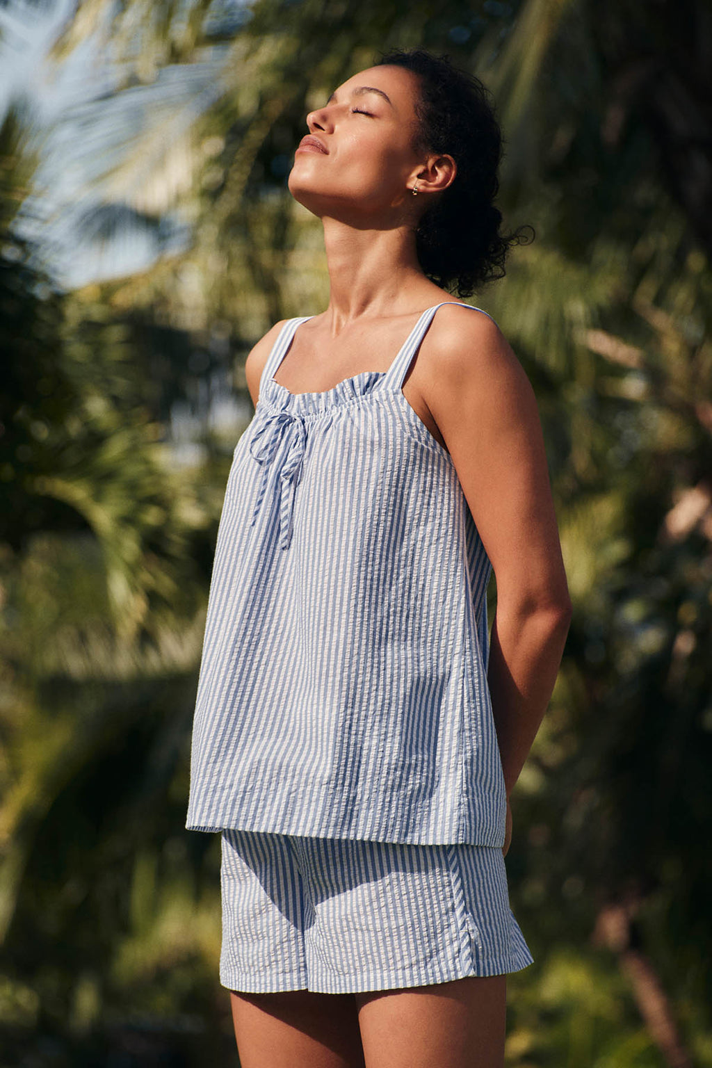 Wearing the Alice Shorts Set in Blue Seersucker, a woman stands outdoors with eyes closed, face turned to the sun and surrounded by lush greenery. This women
