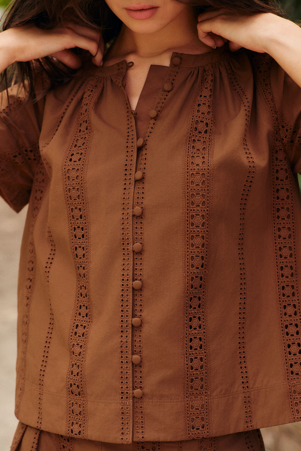 A woman wears the Cafe Shorts Set in Cinnamon Eyelet—a brown, short-sleeved 100% cotton blouse with vertical eyelet lace and fabric-covered buttons. Only her upper torso and part of her face are visible.