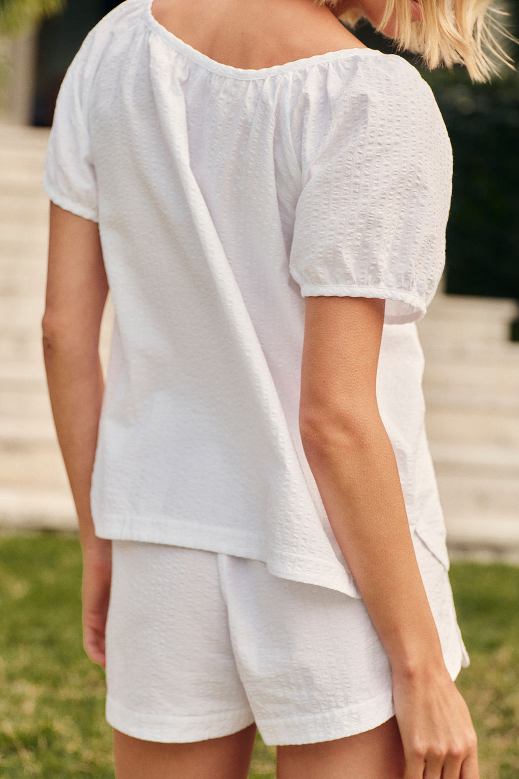 A woman, her back to the camera, stands outdoors on grass with stone steps behind her, wearing the Charlie Shorts Set in White Seersucker.