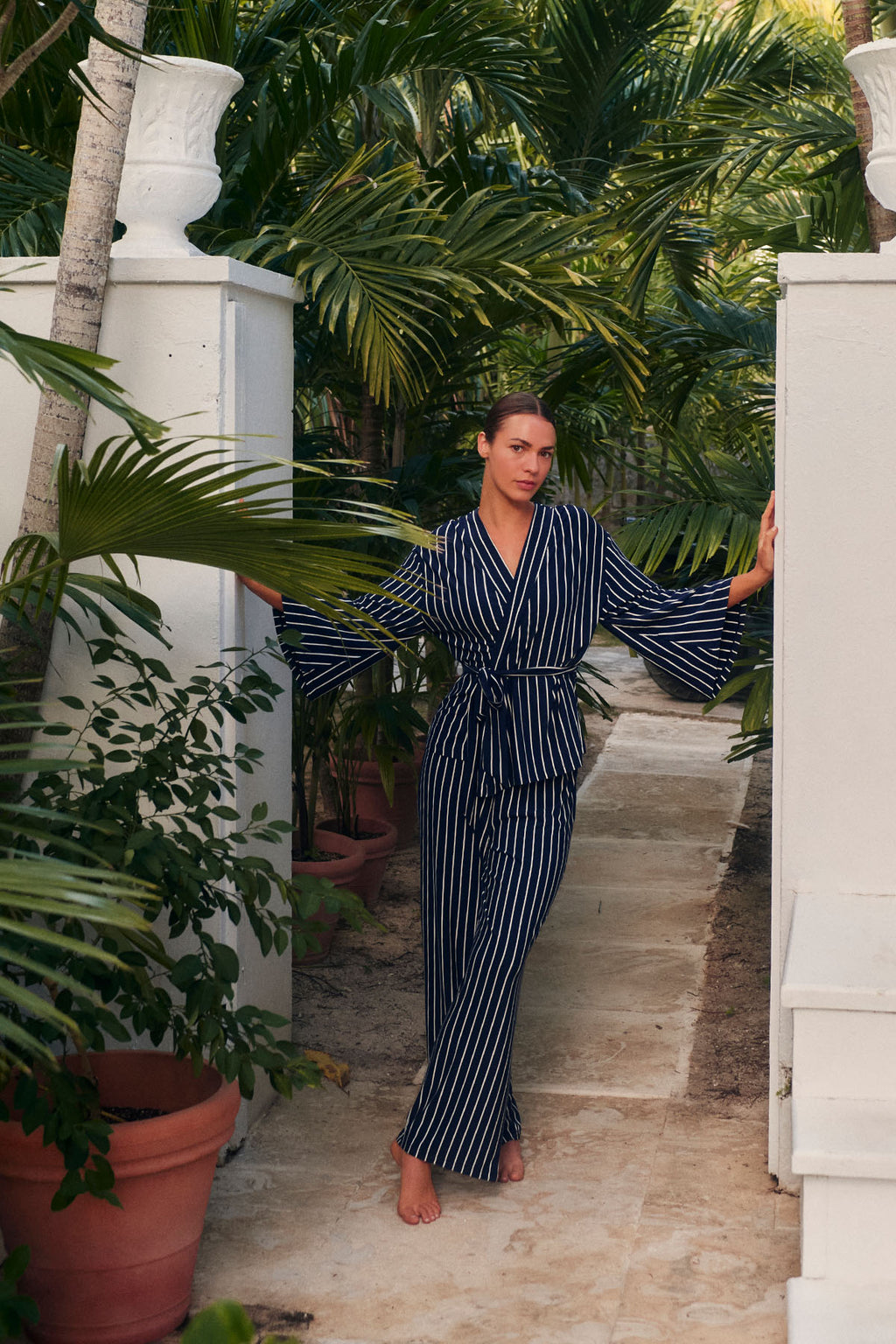 A woman wearing the DreamModal™ Kimono Pajama Set in English Navy Vertical Stripe stands on a stone path between tall tropical plants and white walls, sunlight filtering through the leaves.