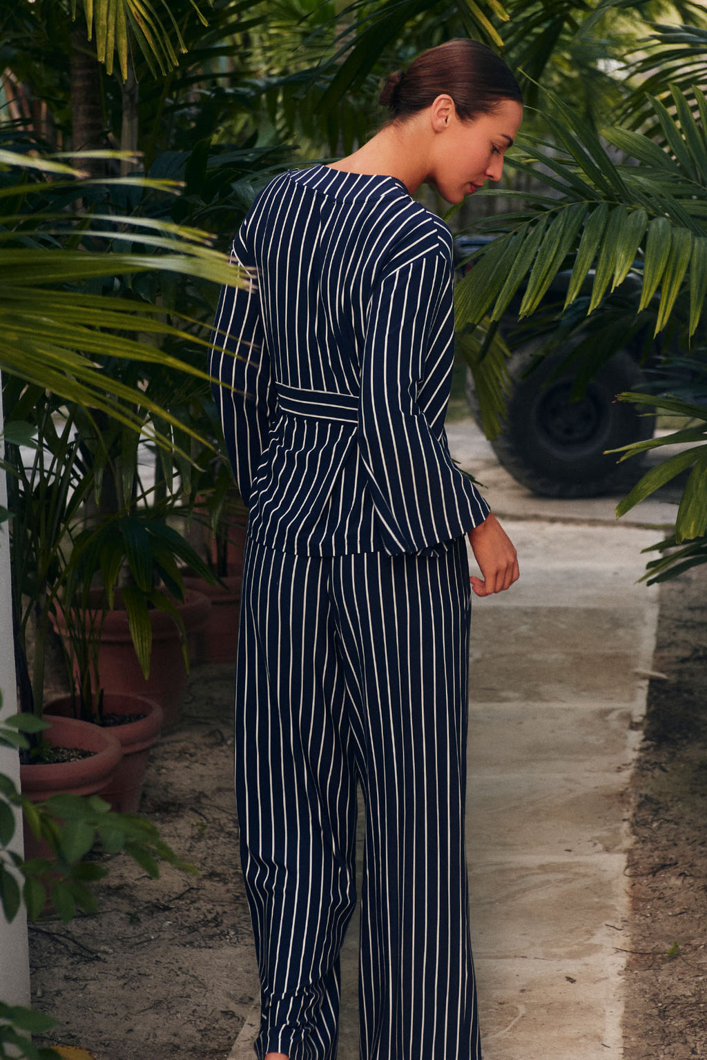 A woman stands on a path surrounded by green plants, wearing the DreamModal™ Kimono Pajama Set in English Navy Vertical Stripe, looking down over her shoulder.