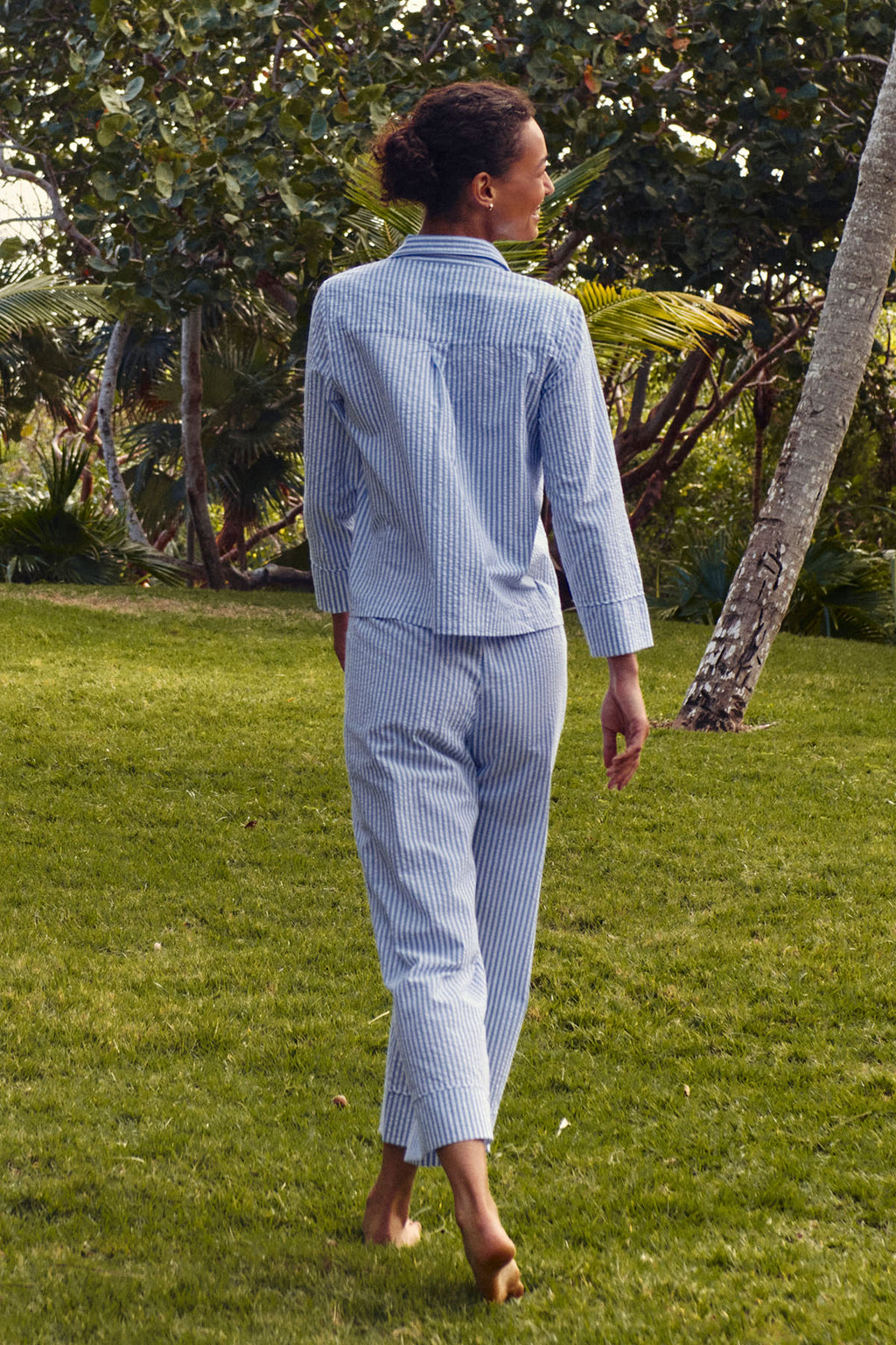 A person wearing the Georgie Pocket Pants Set in Blue Seersucker with a Peter Pan collar walks on grass, surrounded by trees and greenery, seen from behind.