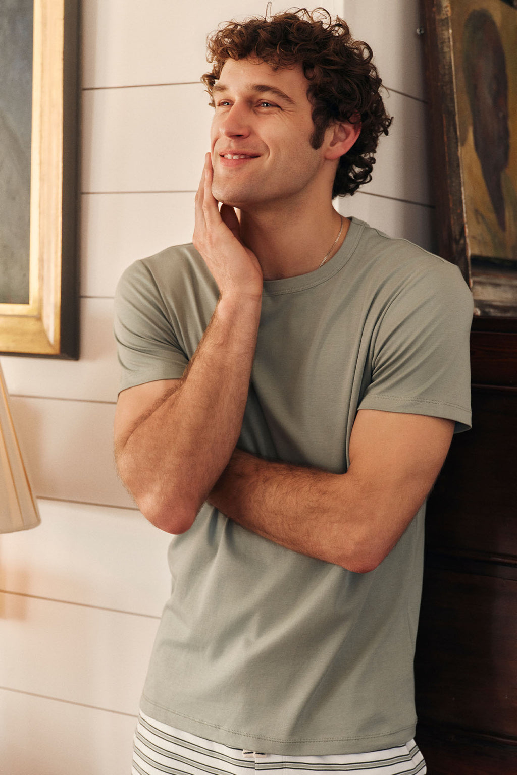 A young man smiles, resting his hand on his face, wearing the Men