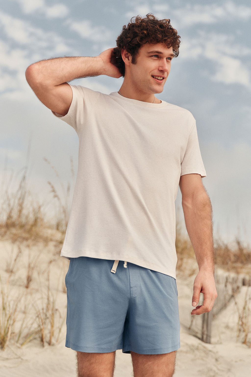 A young man smiles on a sandy beach, wearing the Men