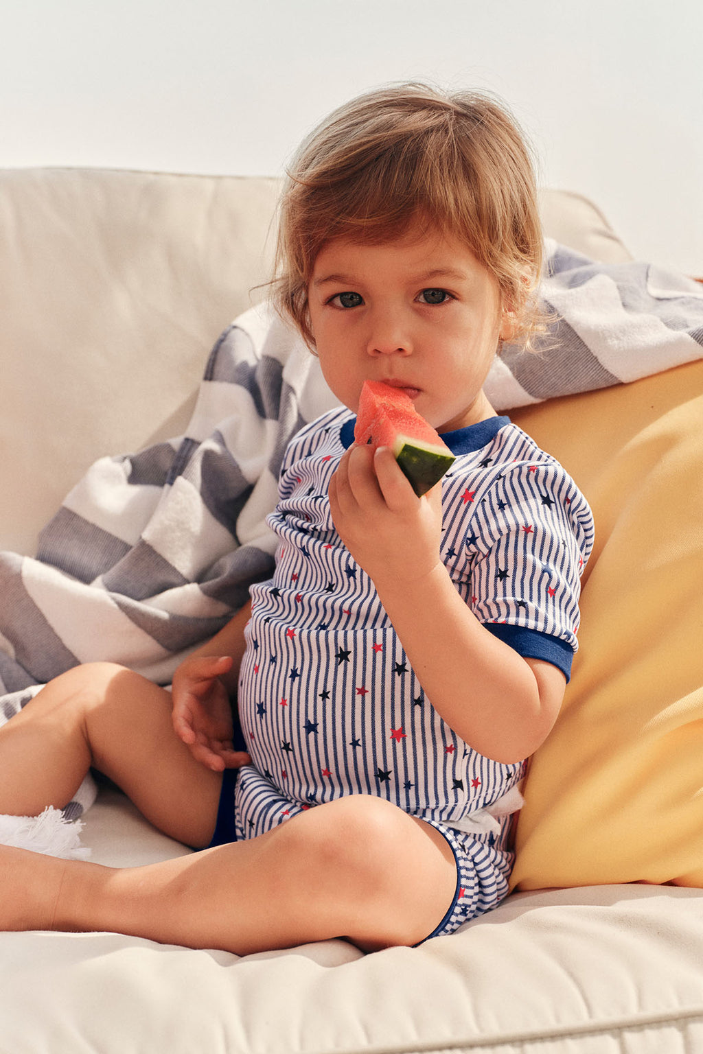 Toddler wearing striped shorts set with blue stars and red accents, sitting on a couch.