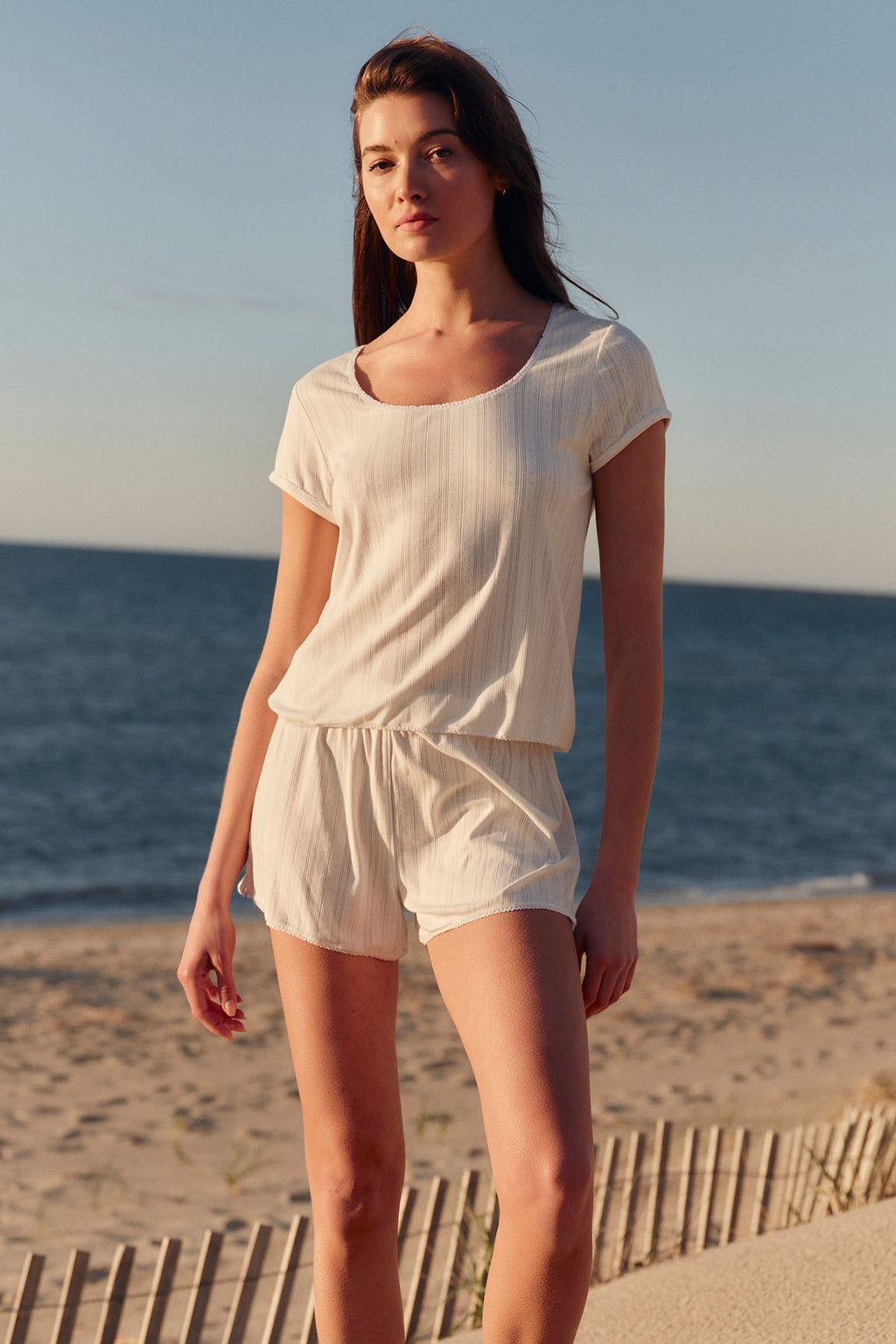 Woman wearing white pointelle short-sleeve top and matching shorts with scalloped hem on a beach.