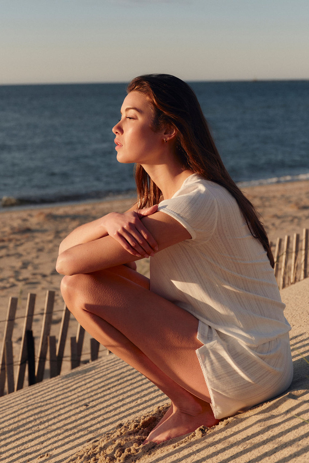 Woman wearing white pointelle shorts set crouching on beach boardwalk with ocean in background.
