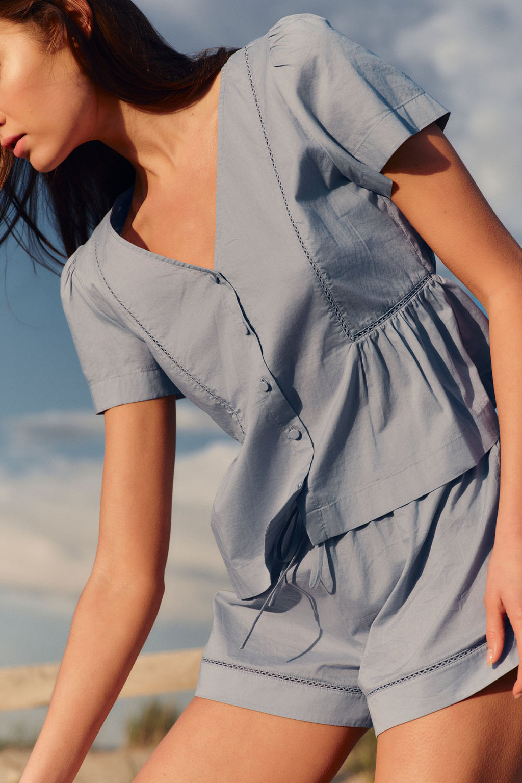 Woman wearing a dusty blue short-sleeve top and matching shorts with button and chain trim details.
