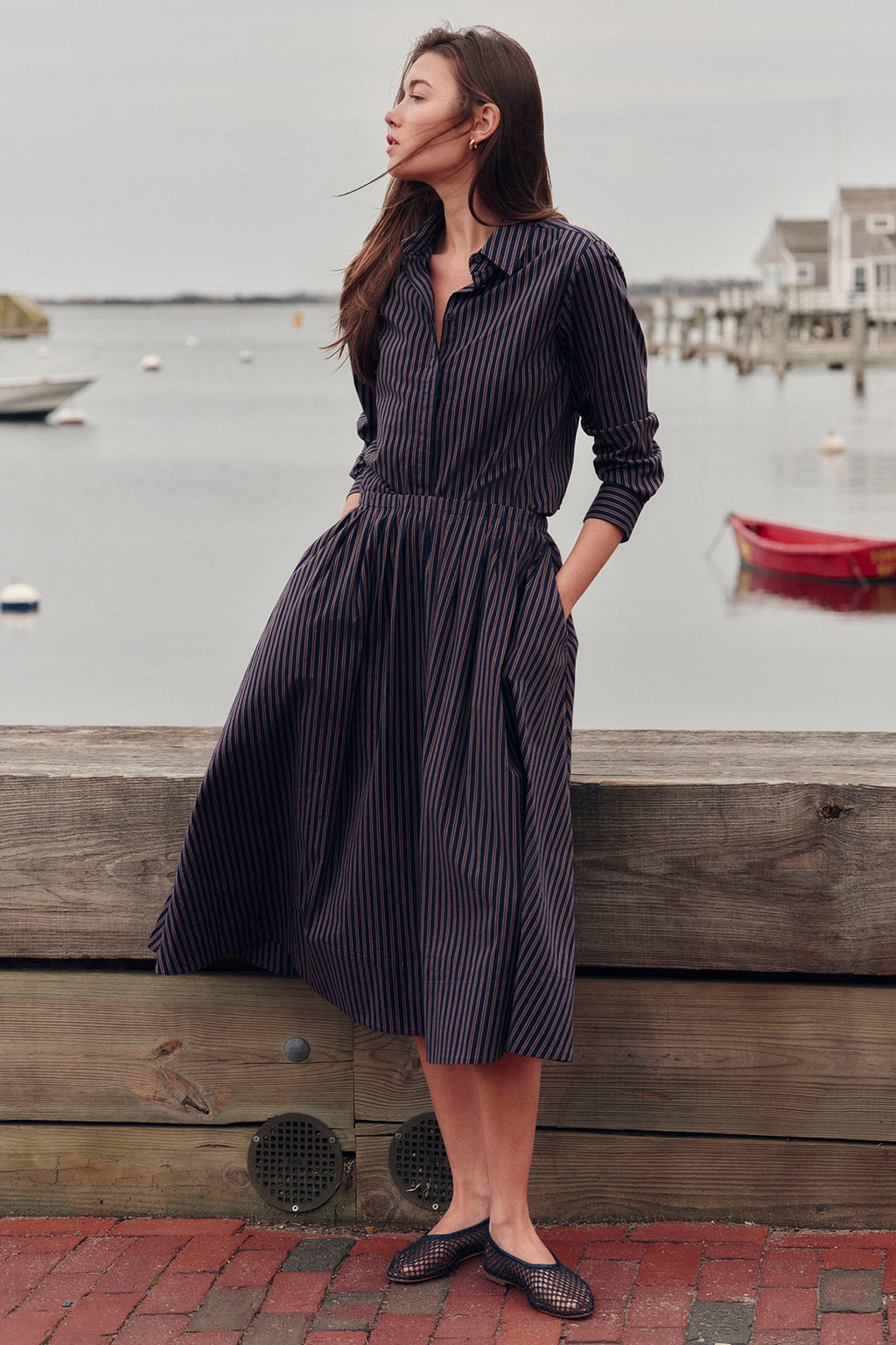 Woman wearing navy striped shirt and matching midi skirt with pockets, standing by waterfront.