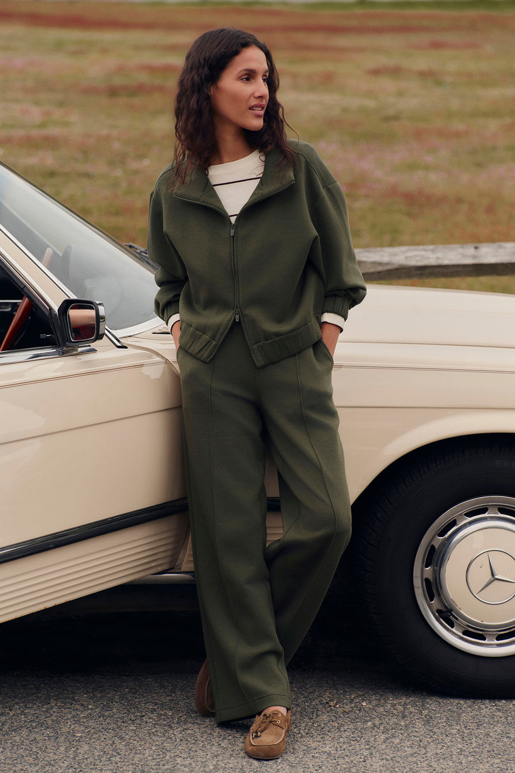 Woman wearing cedar green zip-up jacket and matching pants with white undershirt and brown loafers.