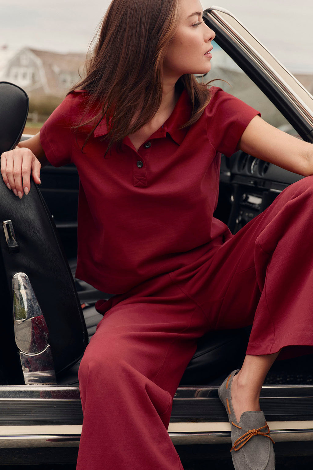 Woman wearing deep red short-sleeve collared shirt and matching pants, seated in convertible car.