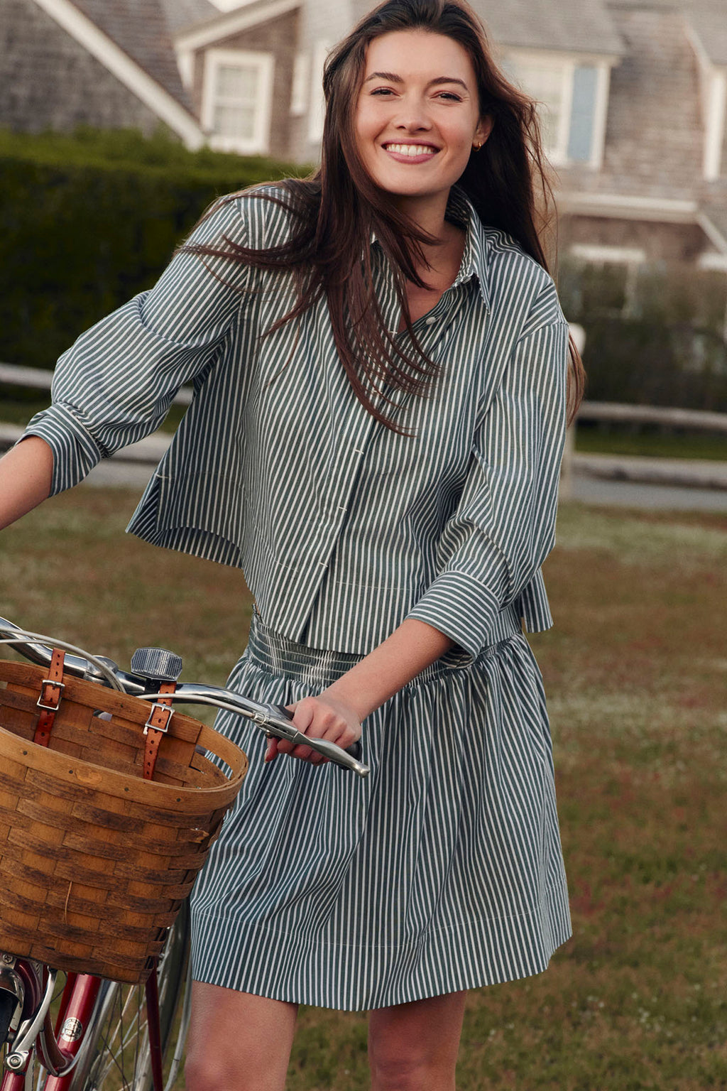 Woman wearing dark green and white striped button-up shirt and matching knee-length skirt, standing with vintage bicycle.