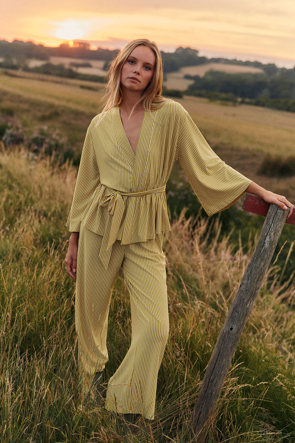 Woman wearing yellow and white striped kimono robe and matching pants in rural field at sunset.