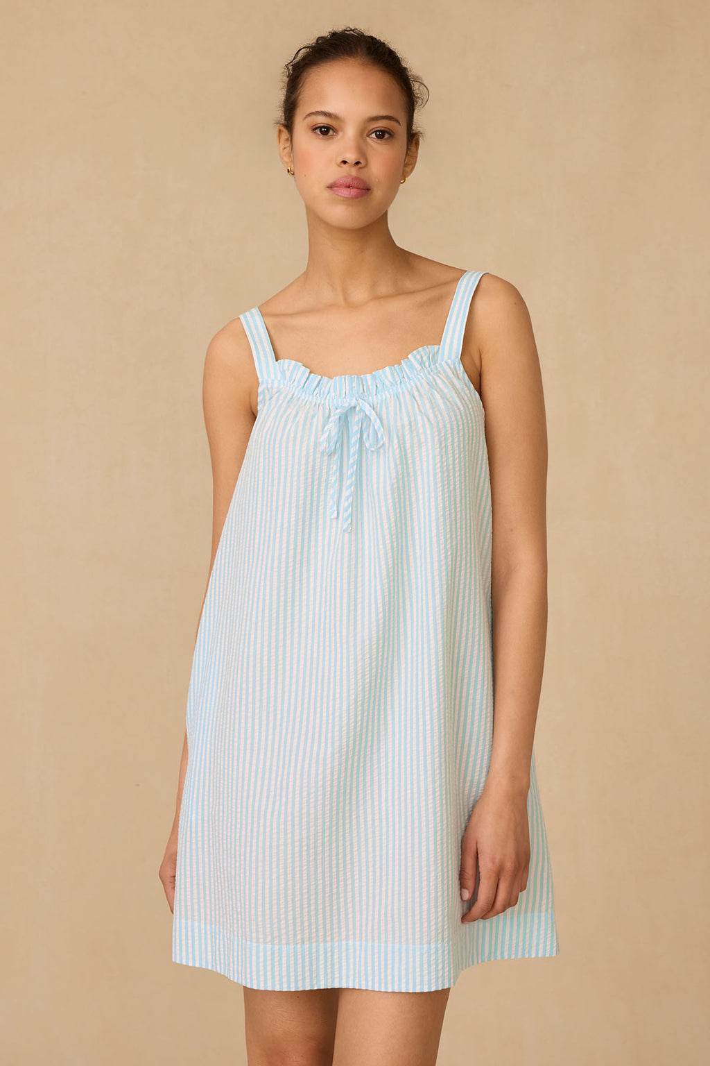 A woman stands against a beige backdrop in the Alice Nightgown in Salt Spray Seersucker, a loose, sleeveless style featuring a bow at the neckline. She gazes at the camera with a neutral expression.