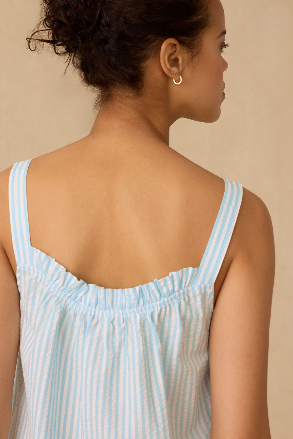 A woman with curly hair in a loose updo, gold hoop earrings, and the Alice Nightgown in Salt Spray Seersucker is shown from the back looking right against a neutral background.