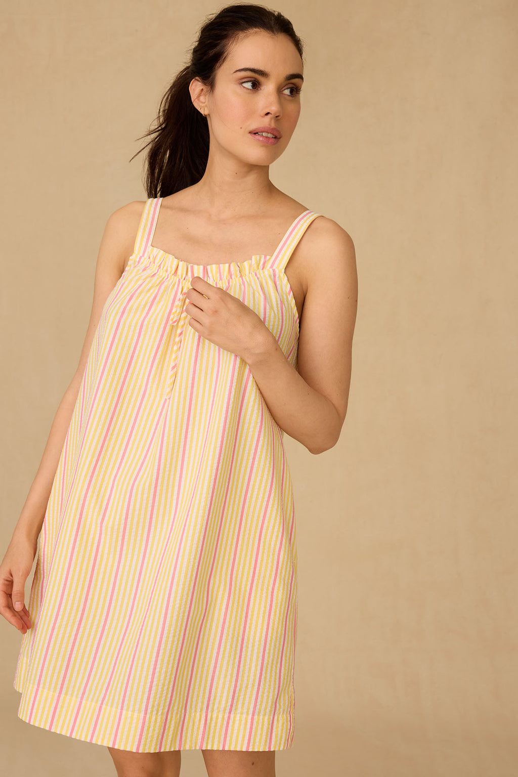 A woman in the Alice Nightgown in Zest Coastline Seersucker stands against a plain beige background, gazing to the side with one hand gently touching her loose-fitting, sleeveless striped dress.