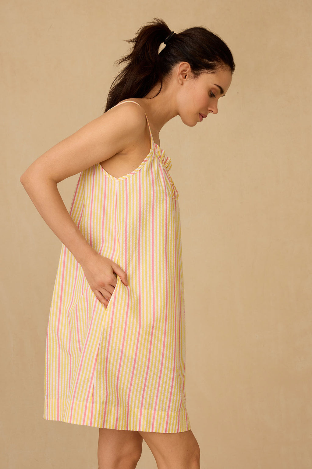 A woman in a ponytail models the sleeveless, loose-fitting Alice Nightgown in Zest Coastline Seersucker, standing in profile against a beige background.