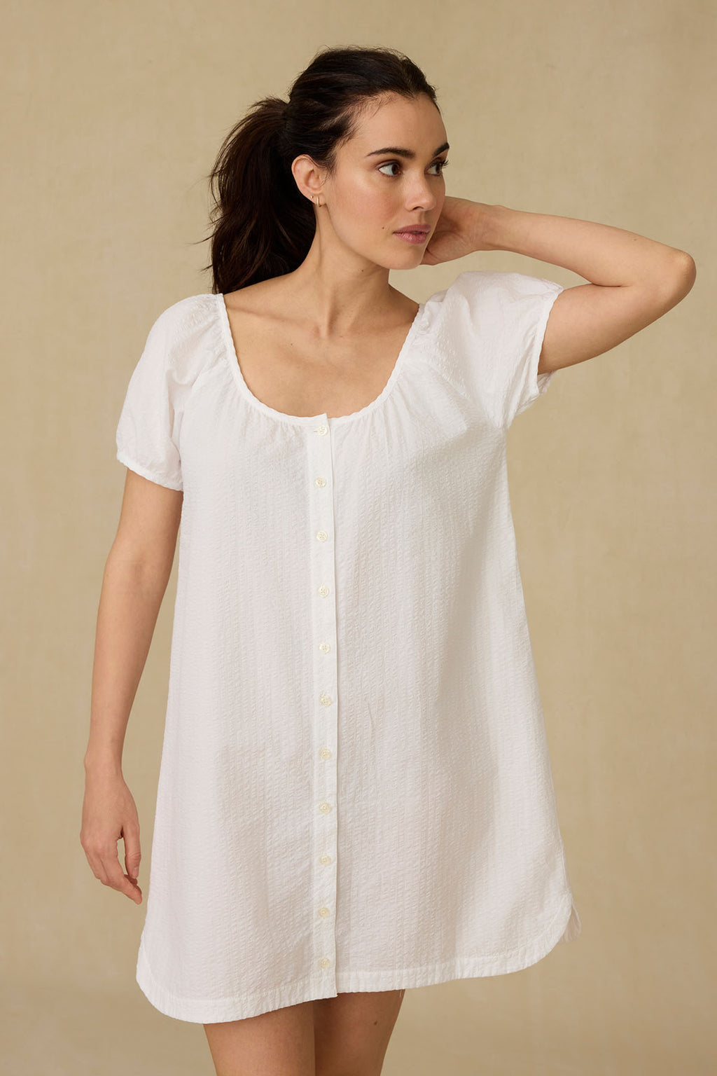 A woman wearing the Charlie Nightgown in White Seersucker, featuring loose short sleeves and a button placket, stands against a plain beige background, gazing to the side.