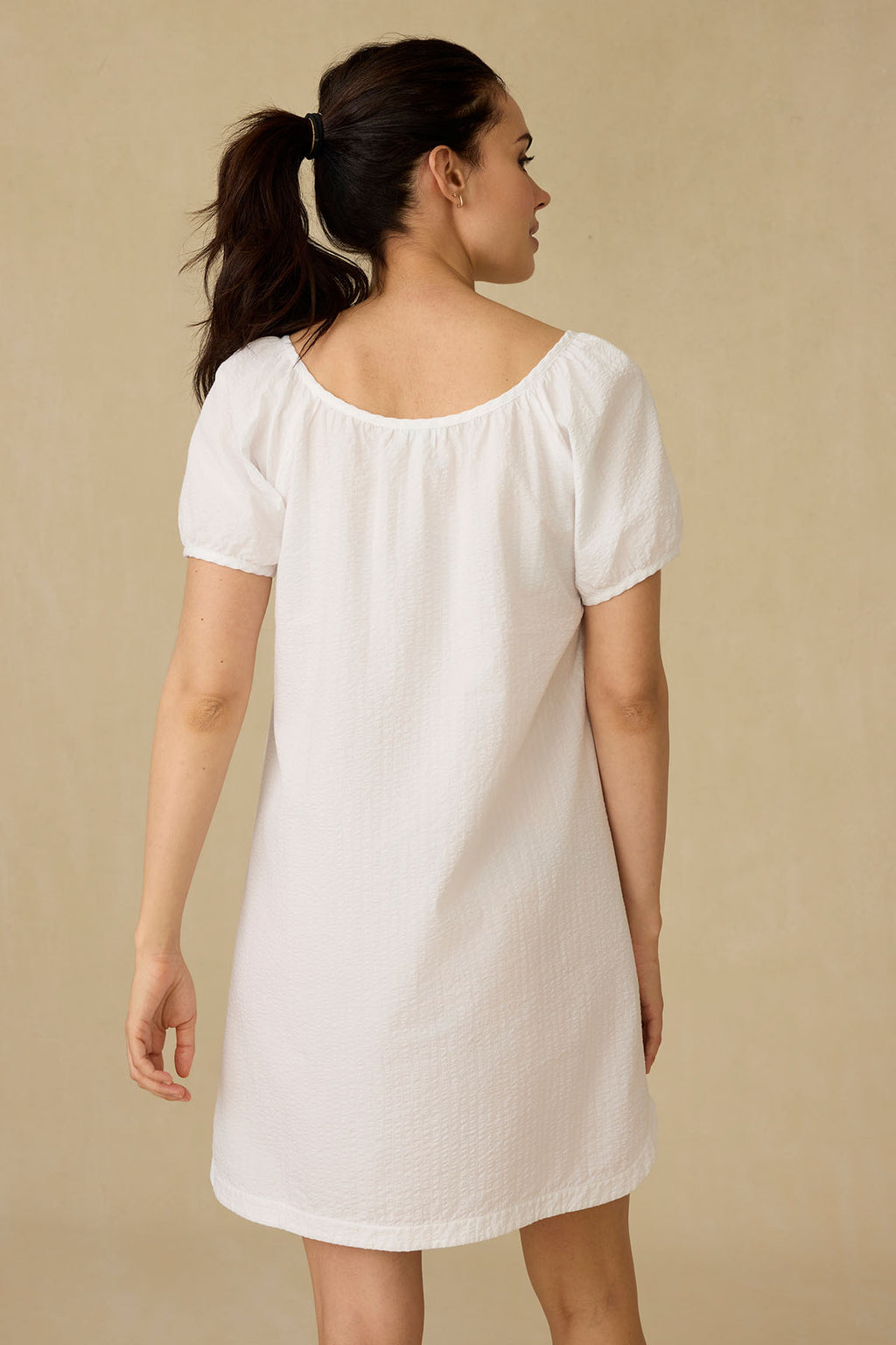 A woman stands with her back to the camera, wearing the Charlie Nightgown in White Seersucker—a short-sleeved, knee-length nightgown with a button placket—against a neutral beige background.