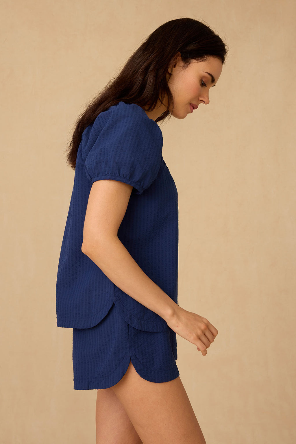 A woman stands in side profile against a beige background, wearing the Charlie Shorts Set in Blue Depths Seersucker, featuring textured Pima cotton and scalloped hems.