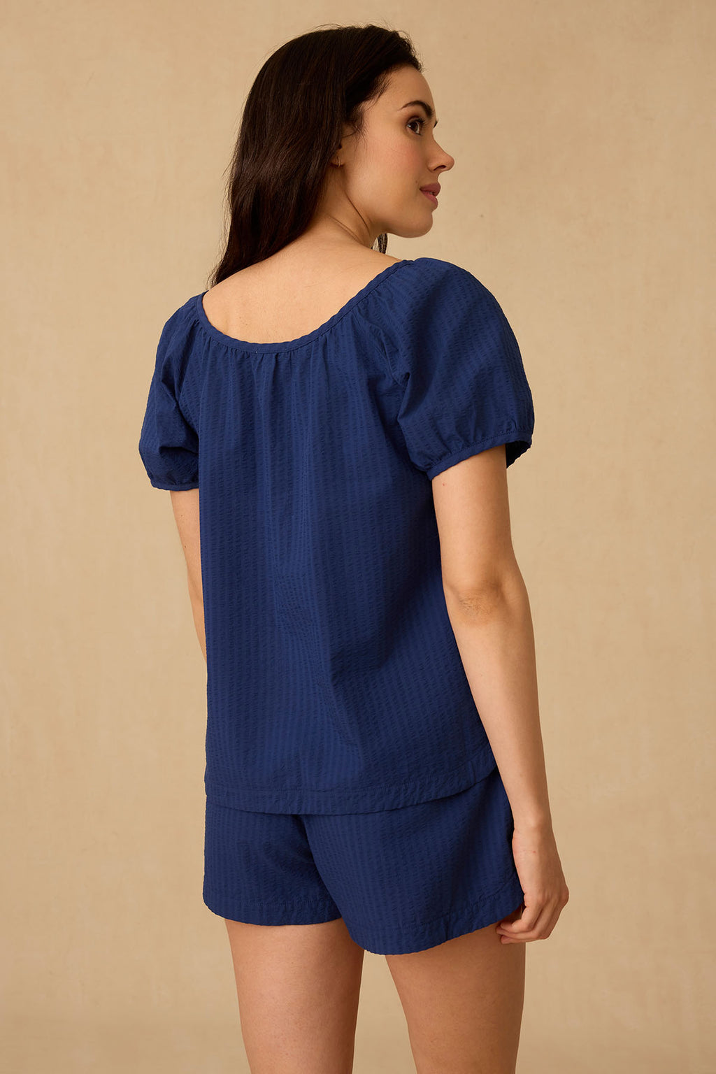 A woman stands facing away, wearing the Charlie Shorts Set in Blue Depths Seersucker—a navy blue short-sleeve top and matching loose-fit shorts in textured Pima cotton seersucker—against a plain beige background.