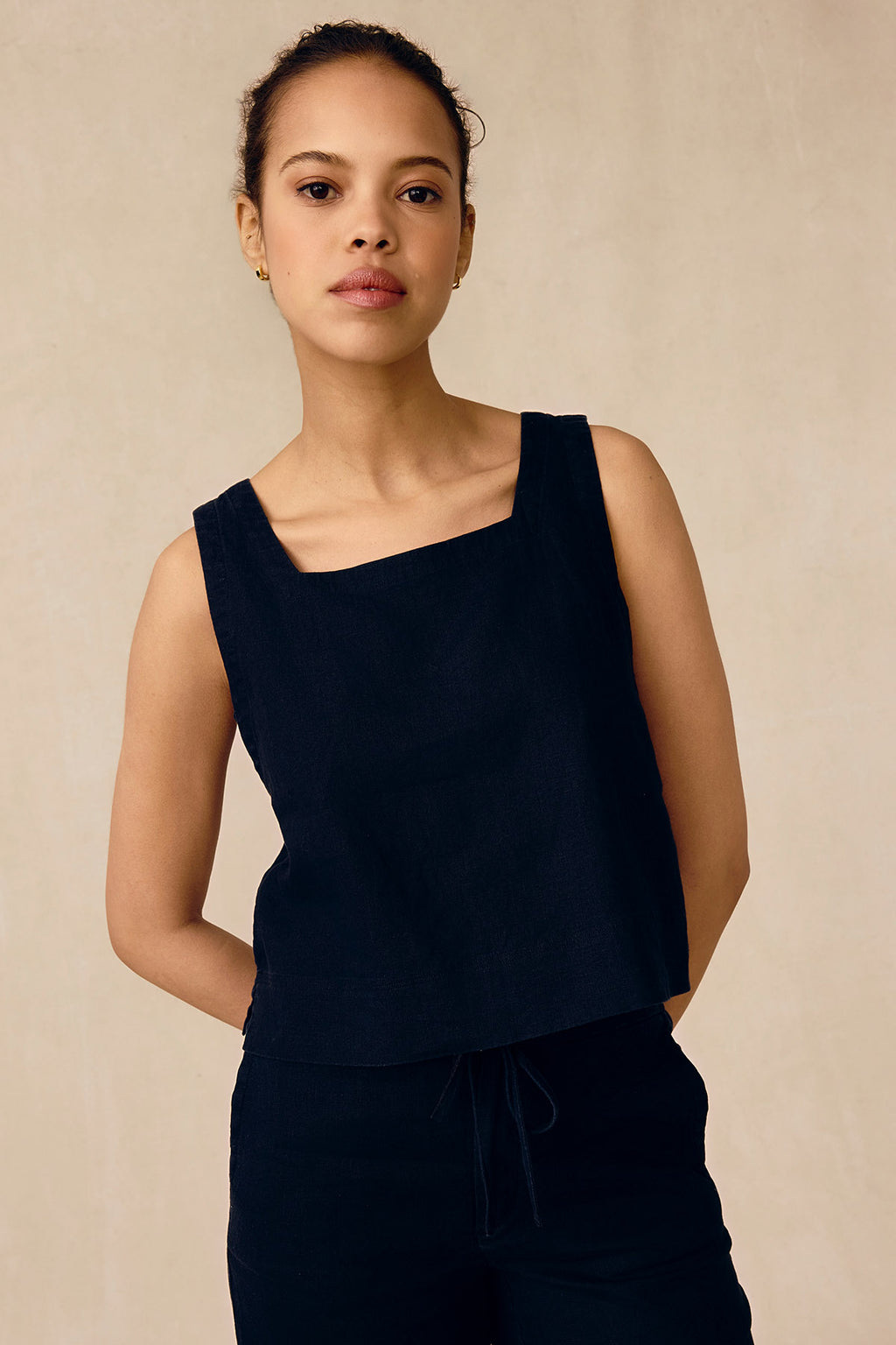 A woman with tied-back hair models the Coupe Pant Set in Black, a sleeveless top and matching linen pants, standing against a plain beige background with her hands behind her back, gazing calmly at the camera.