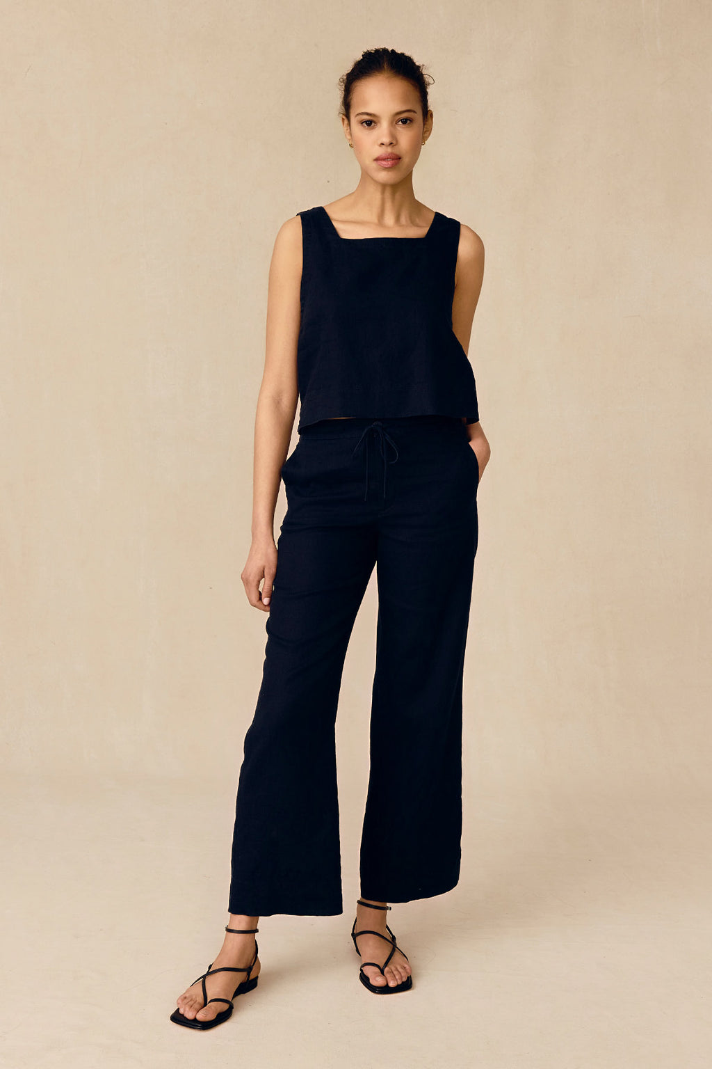 A woman models the Coupe Pant Set in Black, featuring a sleeveless top and wide-leg drawstring pants. She stands against a plain beige background with her hands in her pockets, completing the look with black strappy sandals.