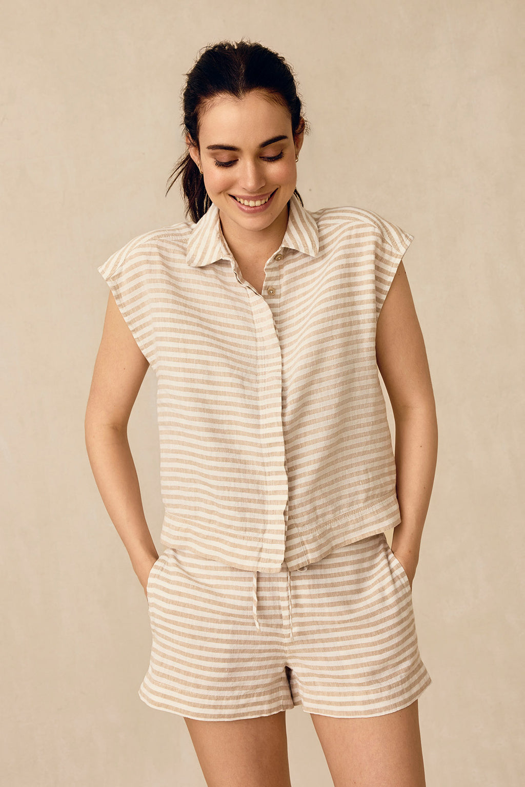 A woman smiles in the Cruise Short Set in Sandstone Amalfi Stripe, featuring a beige and white striped sleeveless button-up shirt and matching shorts—hands in her pockets against a neutral background, perfect for a women’s cruise outfit.