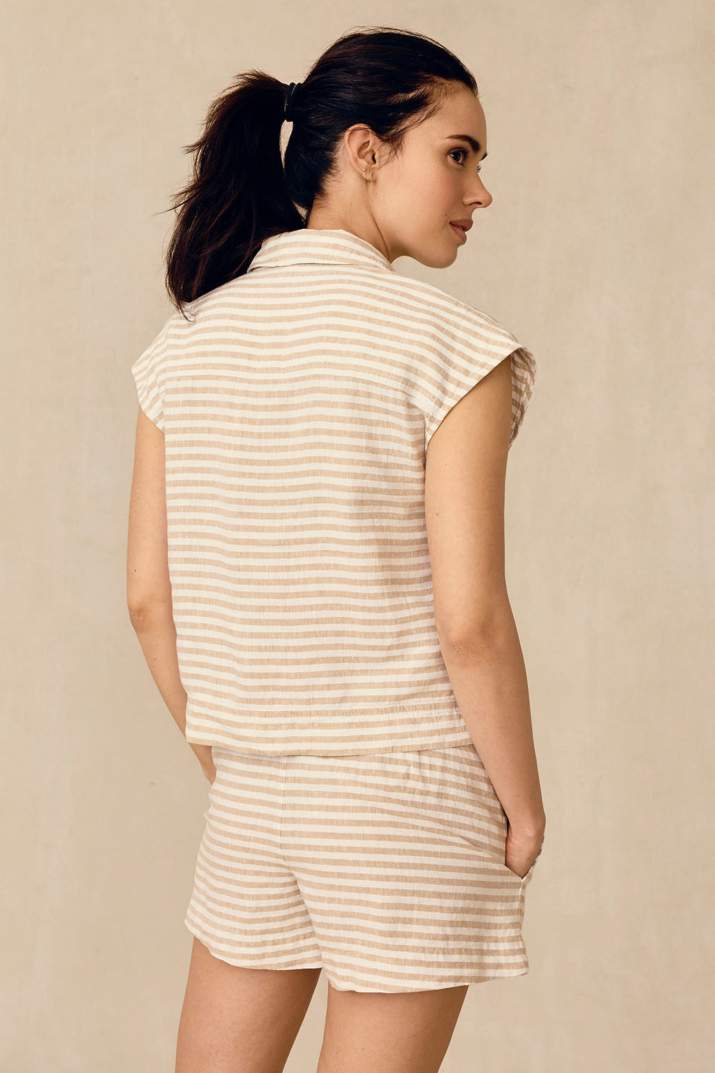 A woman faces away, wearing the Cruise Short Set in Sandstone Amalfi Stripe—short sleeves in beige and white stripes—perfect for a women’s cruise, set against a neutral background.
