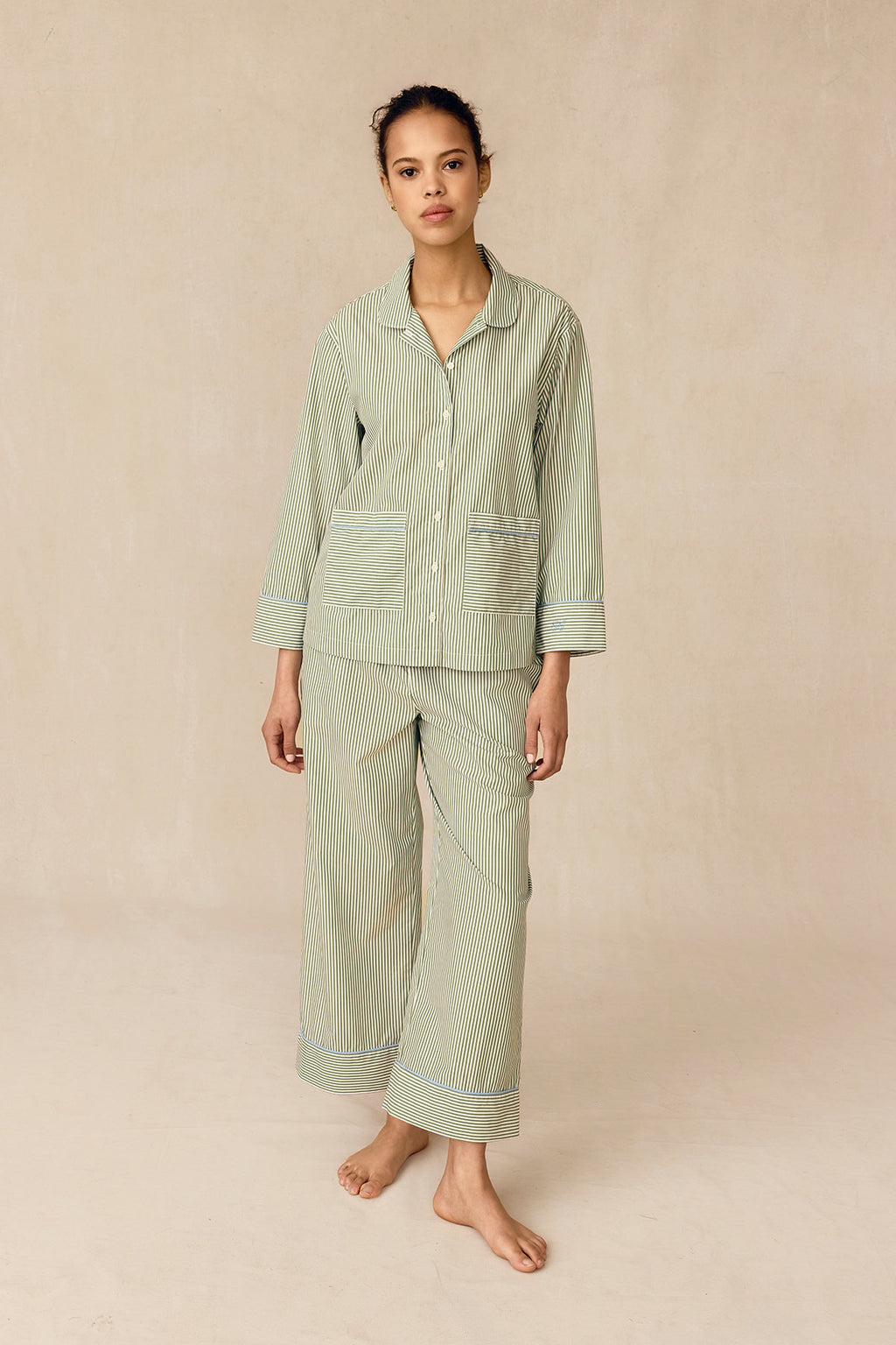 A woman stands against a beige background wearing the Poplin Piped Long-Long Set in Palm Riviera Stripe—a loose-fitting, light green and white striped cotton pajama set with long sleeves, collar, and front pockets. She gazes at the camera neutrally.