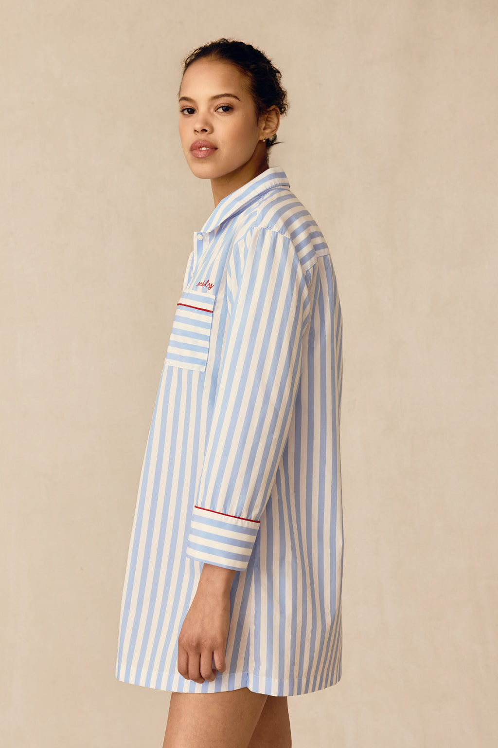 A person stands sideways in the Poplin Piped Nightgown in Baltic Blue Beam Stripe, featuring red trim and a chest pocket, set against a plain beige background.