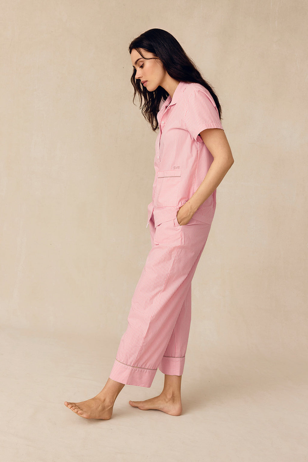 A woman stands on a beige floor, looking down with her hands in her pockets while wearing the Poplin Piped Short-Long Set in Gelato Riviera Stripe—a pink and white striped pajama set with short sleeves and pockets.