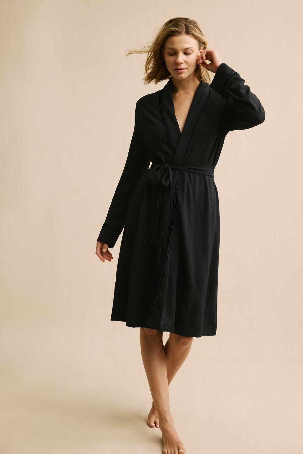 A woman stands against a beige background, wearing the Relax Robe in Black—a knee-length, long-sleeve modal robe with a wrap-around waist tie and a relaxed expression.