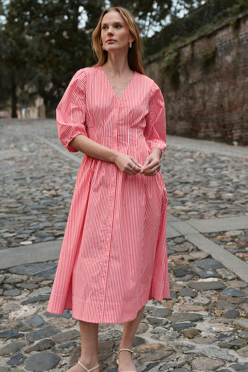 A woman stands on a cobblestone street wearing the Sunday Dress in Tart Bengal Stripe, a red and white cotton poplin midi dress with long puffed sleeves and a V-neck, paired with beige sandals. Trees and a brick wall are visible in the background.
